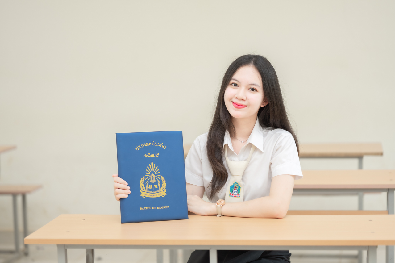 A young woman sitting indoors, holding a graduation certificate proudly.